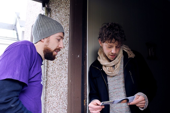 Young man in purple shirt and wool cap talking to a young man at the door.